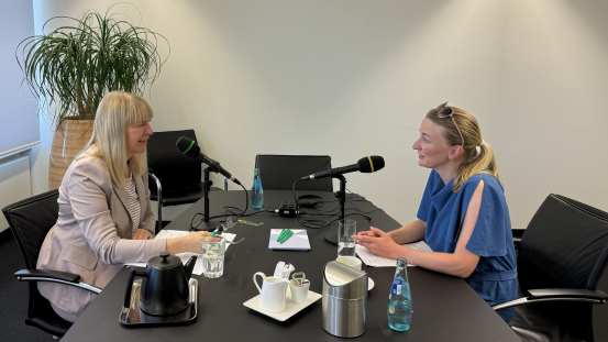 Zwei Frauen sitzen sich an einem Konferenztisch gegenüber und sprechen in Mikrofone. Beide tragen helle Kleidung, eine Frau trägt einen hellen Blazer, die andere ein blaues Oberteil. Auf dem Tisch stehen eine Kaffeekanne, Tassen, ein Glas Wasser, eine Wasserflasche, ein Notizblock und ein Kugelschreiber. Im Hintergrund steht eine große Zimmerpflanze in einem Korb. Die Szene wirkt wie eine Gesprächssituation oder ein Interview.