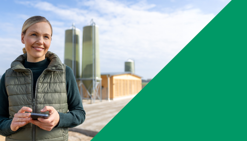 Eine Frau mit blonder Frisur und Zopf trägt eine gesteppte, dunkelgrüne Weste über einem hellen Pullover. Sie hält ein Smartphone in beiden Händen und lächelt. Im Hintergrund stehen moderne landwirtschaftliche Gebäude und Silos vor einem wolkigen Himmel. Die rechte Bildhälfte ist von einer großen, grünen Farbfläche und einer diagonalen Holzstruktur dominiert.